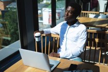 An African American businessman sitting at a table inside a cafe, working on his laptop and thinking, holding a cup of coffee — Stock Photo