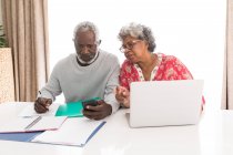 Старшая афроамериканская пара проводит время дома вместе, социальное дистанцирование и самоизоляция в карантинной изоляции во время эпидемии коронавируса, сидя за столом, используя ноутбук, человек, держащий смартфон — стоковое фото