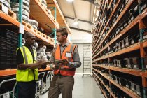 Caucasian and African American male factory workers wearing a high vis vest talking, holding tablet and clipboard. Workers in industry at a factory making hydraulic equipment. — Stock Photo