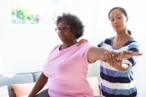 Старша змішана жінка проводить час вдома, відвідуючи змішану расу медсестру, медсестра масажує руку — стокове фото