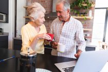 Una pareja de ancianos jubilados caucásicos en casa de pie en una mesa en su cocina, hablando y sonriendo, utilizando un ordenador portátil y beber café juntos, pareja aislante durante coronavirus covid19 pandemia - foto de stock
