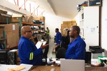 A mixed race male supervisor and an African American male worker in a storage warehouse at a factory making wheelchairs, standing and talking at a workbench — Stock Photo