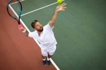 Un hombre de raza mixta que usa blancos de tenis que pasa tiempo en una cancha jugando al tenis en un día soleado, preparándose para golpear una pelota. - foto de stock