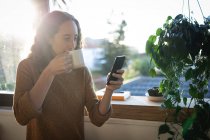 Kaukasische Frau verbringt Zeit zu Hause selbstisolierend und sozial distanziert in Quarantäne während der Coronavirus covid 19 Epidemie, trinkt Kaffee und benutzt ihr Smartphone. — Stockfoto