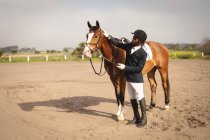 Бічний вид на вправно одягненого афроамериканця, який пестить свого каштанового коня перед стрибком на коні в сонячний день.. — стокове фото