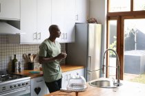Vista lateral de un hombre afroamericano en casa, de pie en la cocina sosteniendo una taza de café y mirando por la ventana al jardín - foto de stock
