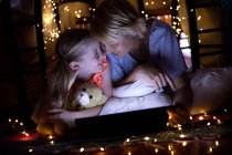 Вид спереди на кавказку, наслаждающуюся семейным времяпрепровождением с дочерью дома вместе, лежащую в палатке, улыбающуюся, с цифровой табличкой, с дочерью, обнимающей своего плюшевого мишку — стоковое фото