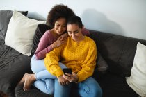 Вид спереду змішаної раси жіноча пара розслабляється вдома, сидячи на дивані і дивлячись на смартфон — стокове фото