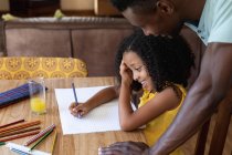Афроамериканська дівчинка в жовтій блузці, соціальна відстань вдома під час карантину, сидячи за столом і малюючи фотографії з татом.. — стокове фото