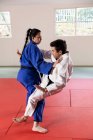 Vue latérale d'un entraîneur de judo masculin de race mixte et d'une judoka féminine de race mixte adolescente, portant du judogi bleu et blanc, pratiquant le judo lors d'un entraînement dans un gymnase. — Photo de stock