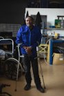 Retrato de un trabajador afroamericano discapacitado de pie usando muletas, usando ropa de trabajo, en un almacén de almacenamiento en una fábrica haciendo sillas de ruedas, mirando a la cámara - foto de stock