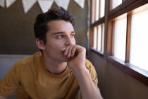 Retrato de un adolescente caucásico sentado en un escritorio en un aula de la escuela mirando por la ventana, pensando - foto de stock