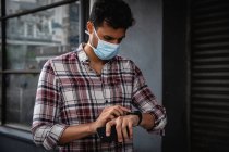 Nahaufnahme eines kaukasischen Mannes mit kariertem Hemd und Gesichtsmaske gegen Luftverschmutzung und Covid19 Coronavirus, der mit seiner Smartwatch durch die Straßen der Stadt läuft. — Stockfoto