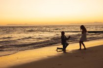 Кавказская пара наслаждается своим временем на пляже во время заката, мужчина стоит на коленях и делает предложение женщине — стоковое фото