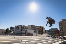 Vista lateral de un hombre caucásico practicando parkour junto al edificio en una ciudad en un día soleado, saltando por encima de las escaleras. - foto de stock