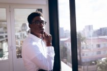 Портрет афроамериканского бизнесмена, одетого в белую рубашку, работающего в современном офисе, стоящего рядом с окном на солнце, трогающего подбородок, смотрящего в камеру и думающего: — стоковое фото