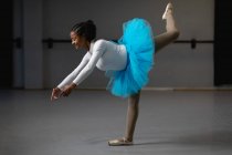 Vista laterale di una ballerina di razza mista con tricot bianco e tutù blu, che balla in uno studio luminoso, alza la gamba e sorride. — Foto stock