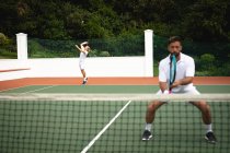 Ein kaukasischer und ein gemischter Rasse Männer in weißen Tennisanzügen verbringen Zeit auf einem Tennisplatz zusammen, spielen Tennis an einem sonnigen Tag, halten Tennisschläger, einer von ihnen schlägt einen Ball mit einem Schläger — Stockfoto