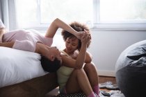 Front view close up of a mixed race female couple relaxing at home in the bedroom in the morning, one lying on her back on the bed and the other sitting beside her on the floor, smiling at each other and holding hands — Stock Photo