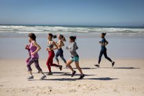 Vista lateral de un grupo multiétnico de amigas disfrutando de hacer ejercicio en una playa en un día soleado, corriendo a la orilla del mar. - foto de stock