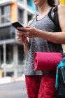 Front view mid section of a fit Caucasian woman on her way to fitness training on a cloudy day, carrying sports bag and a yoga mat, standing in the street, using her smartphone and wearing earphones — Stock Photo