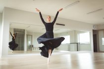 Ballerina di danza femminile attraente caucasica con balletto rosso per capelli, indossa un abito nero e lungo, si prepara per una lezione di balletto in uno studio luminoso, si concentra sul suo esercizio, sorridendo.. — Foto stock