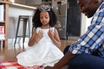 Menina afro-americana vestindo uma coroa de brinquedo, distanciamento social em casa durante o confinamento de quarentena, brincando com seu pai, tendo uma festa de chá de boneca . — Fotografia de Stock
