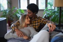 Vue de face gros plan d'un jeune homme métis et d'une jeune femme caucasienne profitant du temps passé à la maison, assis dans leur salon et embrassant, l'homme embrasse la femme sur son front. — Photo de stock