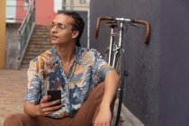 Vue de face rapprochée d'un homme de race mixte avec de longs dreadlocks dehors et autour dans la ville par une journée ensoleillée, assis dans la rue, en utilisant un smartphone, avec son vélo appuyé contre le mur à côté de lui. — Photo de stock