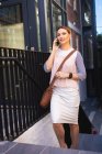 Una mujer de negocios caucásica en movimiento en un día soleado, caminando por las escaleras, hablando en su teléfono inteligente y usando un reloj inteligente - foto de stock