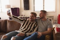 Пара белых мужчин, отдыхающих дома, сидящих на диване, обнимающихся, улыбающихся и делающих селфи со своим смартфоном — стоковое фото