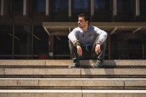 Vue de face d'un homme caucasien pratiquant le parkour près de l'immeuble dans une ville par une journée ensoleillée, se reposant prendre une pause, assis sur les escaliers. — Photo de stock