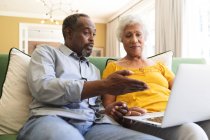 Завершити щасливу подружню пару афроамериканців на пенсії, які сидять вдома на дивані у своїй вітальні, спілкуються та користуються портативним комп 