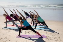 Visão traseira de um grupo multi-étnico de amigas que gostam de se exercitar em uma praia em um dia ensolarado, praticando ioga, de pé na posição de ioga. — Fotografia de Stock