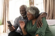 A senior African American couple spending time at home together, the man holding a smartphone and taking a selfie, making a video call to friends or relatives, waving — Stock Photo