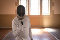 African American sportsman wearing protective fencing outfit during a fencing training session, preparing for a duel, holding an epee in front of face. Fencers training at a gym. — Stock Photo