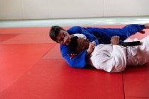 Vue latérale d'un entraîneur de judo masculin de race mixte et d'un judoka masculin de race mixte adolescent portant du judogi bleu et blanc, pratiquant le judo lors d'un entraînement dans une salle de gym. — Photo de stock