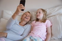 Front view of a Caucasian woman enjoying family time with her daughter at home together, lying on bed in their bedroom, smiling and taking selfies with her smartphone — Stock Photo