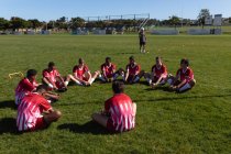 Frontansicht einer multiethnischen Männermannschaft mit Rugby-Spielern im Teamdress, die auf dem Spielfeld sitzt und sich mit ihrem Trainer im Hintergrund aufwärmt. — Stockfoto