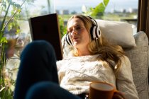 Vista frontal de perto de uma mulher caucasiana sentada em sua sala de estar na frente de uma janela em um dia ensolarado, lendo um livro e usando fones de ouvido — Fotografia de Stock