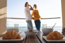 Frühstück auf einem Tisch liegend mit einem kaukasischen Paar, das auf einem Balkon steht und sich im Hintergrund umarmt. Soziale Distanzierung und Selbstisolierung in Quarantäne. — Stockfoto