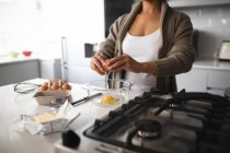 Вид спереду середня секція жінки вдома, стоячи на кухні біля стільниці, готуючи сніданок, розбиваючи яйце в миску — стокове фото