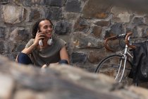 Передний вид смешанного расового человека с длинными дредами в городе в солнечный день, сидящего у стены на улице и улыбающегося, используя смартфон, с велосипедом, прислоненным к стене рядом. — стоковое фото