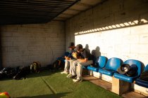 Vue latérale de deux joueurs de baseball de race mixte, lors d'un match de baseball par une journée ensoleillée, assis sur un banc, regardant vers le bas, se préparant à aller sur un terrain — Photo de stock