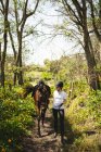 Vista frontal de una jinete caucásica casualmente vestida que conduce a un caballo castaño a través de un camino en el bosque durante un día soleado. - foto de stock