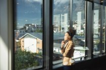 Una mujer de negocios de raza mixta que trabaja en una oficina moderna, mirando por una ventana, tocándose la barbilla y pensando, en un día soleado - foto de stock