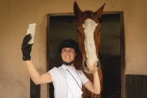 Vista frontale da vicino di una cavallerizza caucasica vestita con disinvoltura usando il suo smartphone e facendo un selfie con il suo cavallo di castagno in piedi in una stalla. — Foto stock