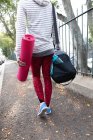 Vista posteriore sezione bassa di una donna in forma sulla strada per l'allenamento fitness in una giornata nuvolosa, portando borsa sportiva e un tappetino yoga — Foto stock