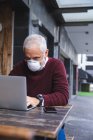 Старший кавказский мужчина сидит за столом на кофейной террасе в маске против коронавируса, ковид 19, используя смартфон и ноутбук. — стоковое фото
