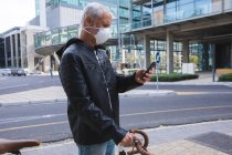 Hombre caucásico mayor por las calles de la ciudad durante el día, usando una máscara facial contra el coronavirus, covid 19, moviendo su bicicleta y usando un teléfono inteligente. - foto de stock